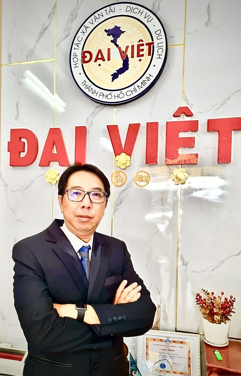 Giang Minh Tuấn mặc vest, khoanh tay đứng trước logo Hợp tác xã Đại Việt